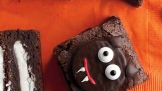 Five decorated brownie squares, some with cookie filling, are topped with chocolate, candy eyes, and icing to resemble vampire faces—perfect spooky Halloween treats arranged on festive orange and black surfaces.