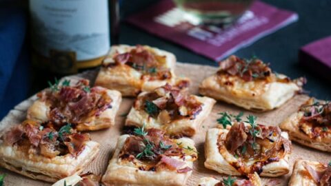 A wooden board with Make Ahead Holiday Appetizers featuring puff pastry topped with herbs, alongside a glass and bottle of white wine on a dark surface.
