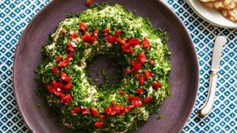 A round cheese and herb spread topped with chopped chives and red peppers is arranged on a purple plate next to a knife and a plate of crackers—a perfect choice for Make Ahead Holiday Appetizers.
