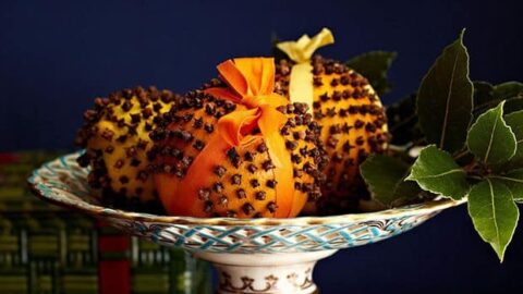 A decorative plate holds orange pomanders—clove-studded oranges—and a drawstring pouch, with holly leaves nearby, set on a wooden surface against a dark blue background.