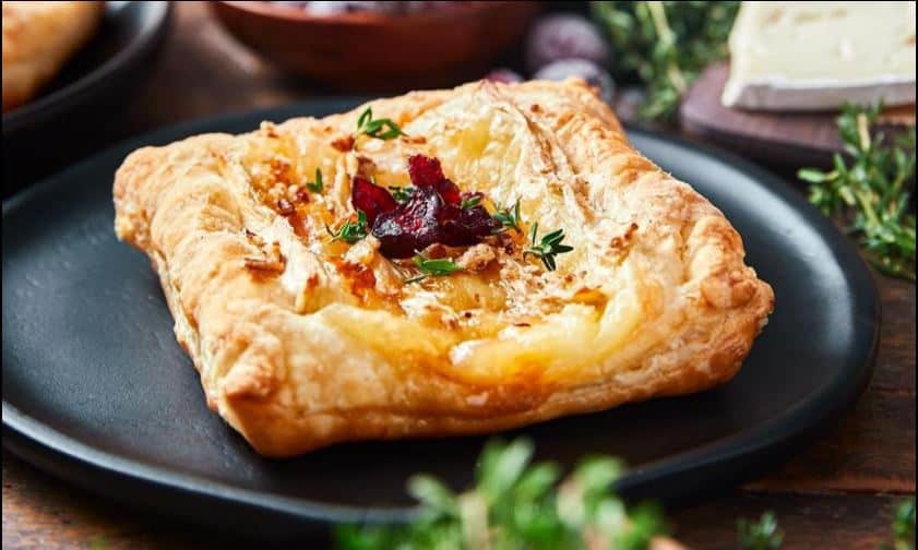 A square puff pastry topped with melted cheese, cranberries, nuts, and herbs sits on a black plate-one of the perfect Easy New Year's Eve Appetizers-surrounded by greenery and cheese in the background.