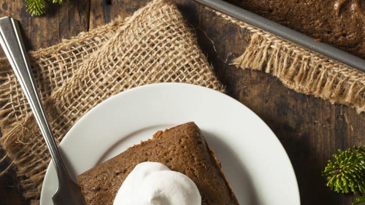A slice of gingerbread cake with whipped cream sits on a white plate, next to a baking pan of gingerbread cake and pine sprigs atop a rustic wooden table.