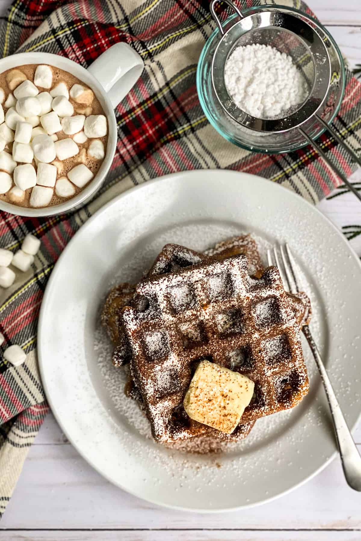 Three gingerbread waffles stacked on a white plate with a pat of butter and dusted with powdered sugar for serving.