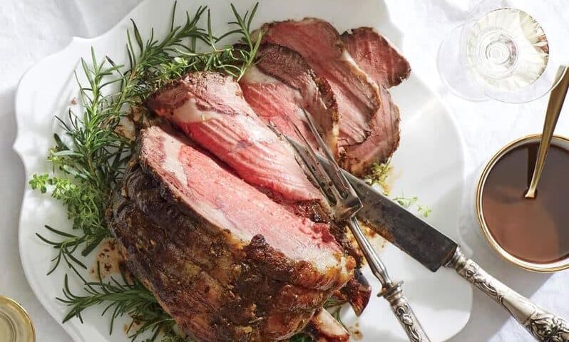 Sliced roast beef on a white platter garnished with fresh herbs, served with a savory gravy sauce and utensils on the side—perfect for your Christmas dinner celebration.