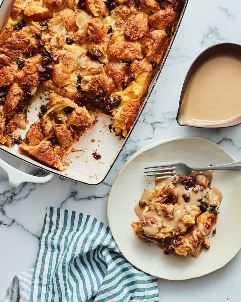 A baking dish with chocolate chip bread pudding, perfect for Christmas breakfast, a slice served on a plate with sauce poured over it, a sauce boat, and a striped napkin on a marble surface.