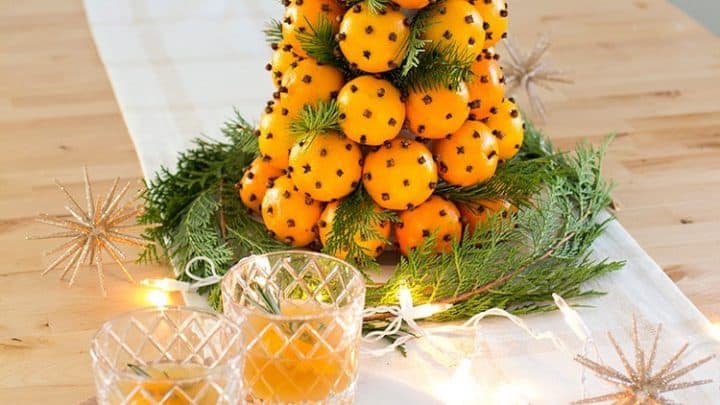 Orange pomanders made of oranges and cloves form a striking centerpiece with greenery on a wooden table, surrounded by cheese, olives, and two glasses of orange-colored drinks.