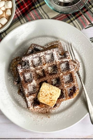 Gingerbread waffles on a plate topped with butter and powdered sugar with a mug of hot chocolate nearby.
