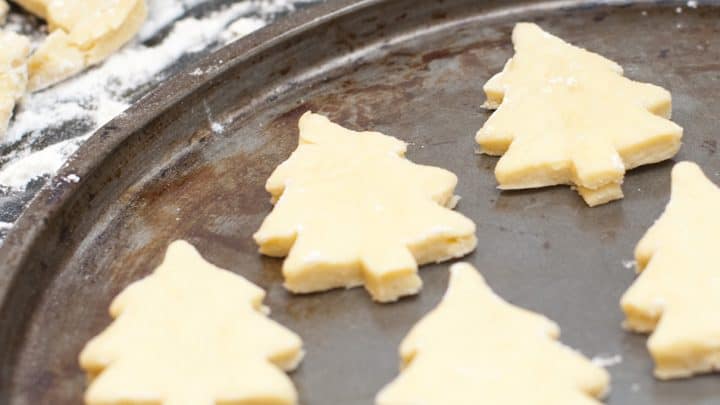 Unbaked Christmas tree-shaped cookie dough, perfect for Christmas cookies, sits on a baking sheet with a rolling pin and dough scraps in the background.
