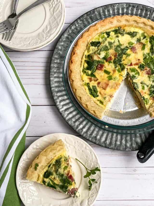Slice of quiche on a white plate with the whole quiche in a metal pie dish in the background.