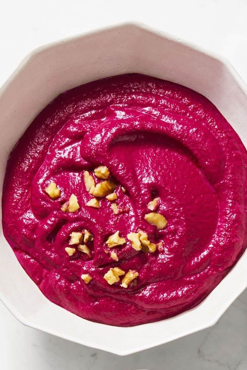 A white bowl filled with bright pink beet hummus, topped with chopped walnuts, on a white surface-one of the perfect easy New Year's Eve appetizers to delight your guests.