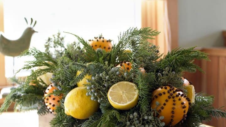 A wooden box centerpiece with orange pomanders, whole and halved lemons, green foliage, and small berries sits on a wooden table near a window.