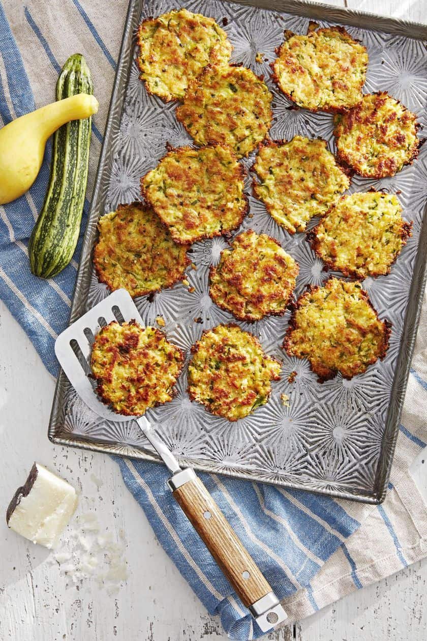 Baked zucchini fritters arranged on a patterned baking sheet with a spatula, next to two raw squash and a wedge of cheese on a striped cloth-perfect as Easy New Year's Eve Appetizers.