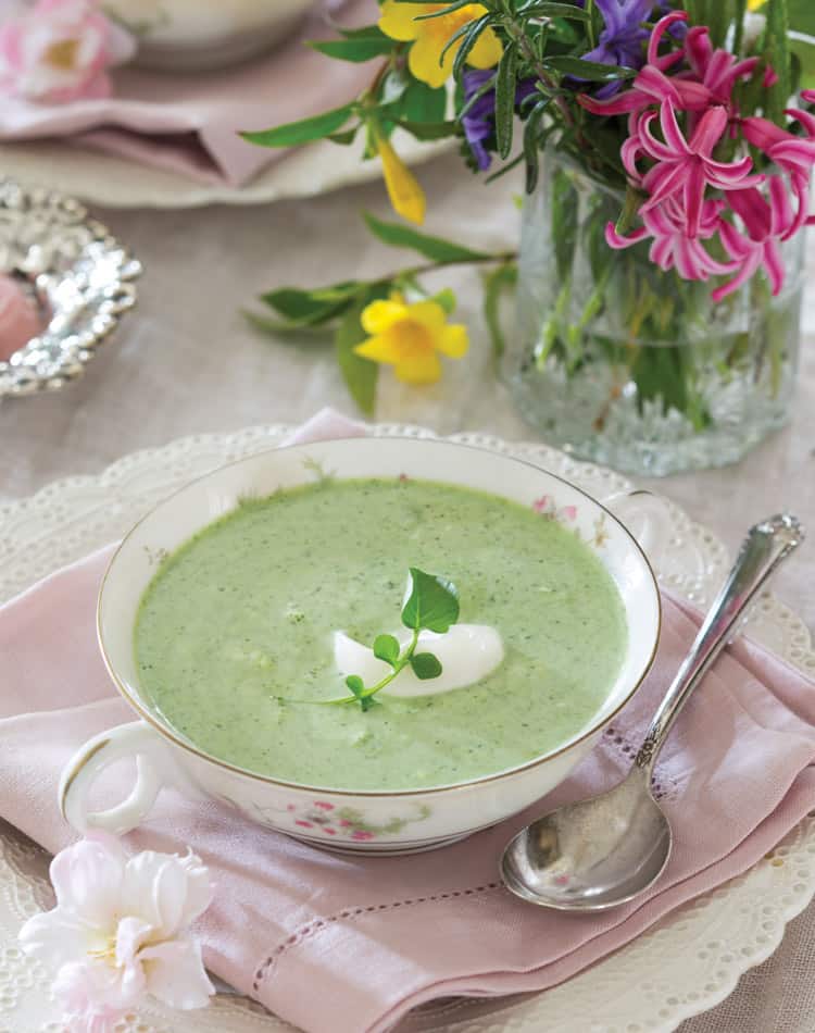 A bowl of green soup with a dollop of cream and herb garnish, set on a pink napkin beside a spoon and a vase of colorful flowers-perfect for a cozy Valentine's Day Afternoon Tea.