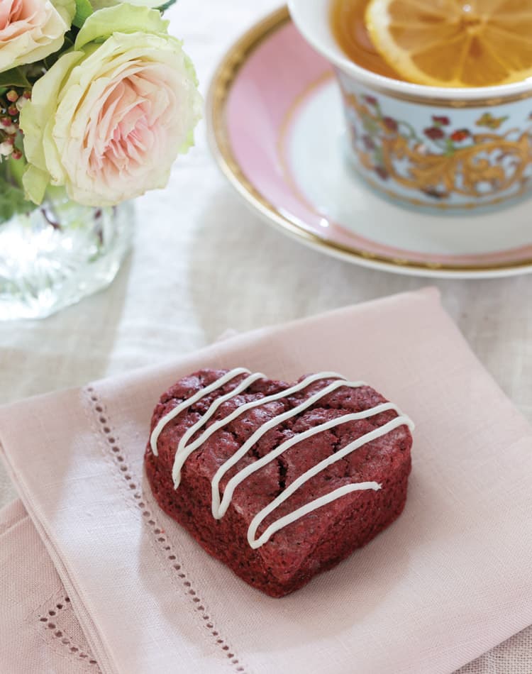 Heart-shaped red velvet cake with white icing drizzle on a pink napkin, perfect for a Valentine's Day Afternoon Tea, served next to a cup of tea with lemon slice and a vase of flowers.