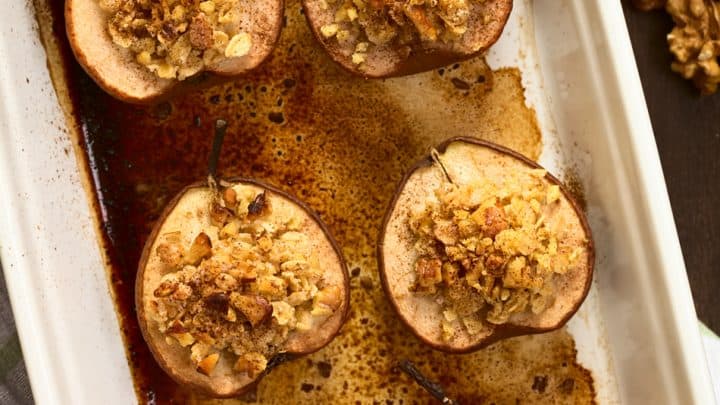 Baked pear halves grace a rectangular dish, each crowned with a blend of walnuts and oats. Caramelized juices surround the baked pears.