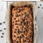 A loaf of chocolate chip banana bread in a parchment-lined pan, topped with extra chocolate chips, rests on a wire rack with scattered chocolate chips nearby.