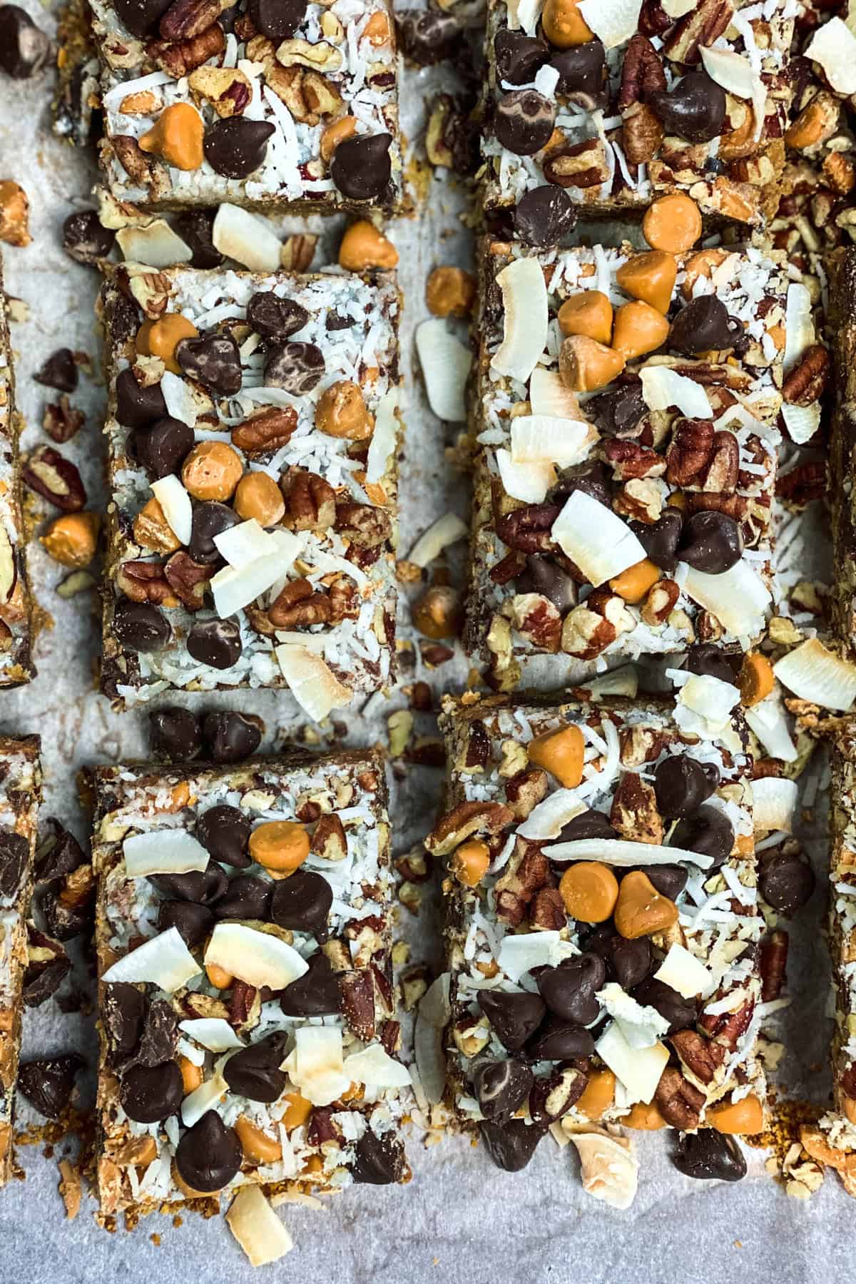Close-up of several Magic Cookie Bars on parchment paper. Topped with chocolate chips, butterscotch chips, pecans, coconut flakes, and banana chips, these delightful treats boast a textured and colorful appearance.
