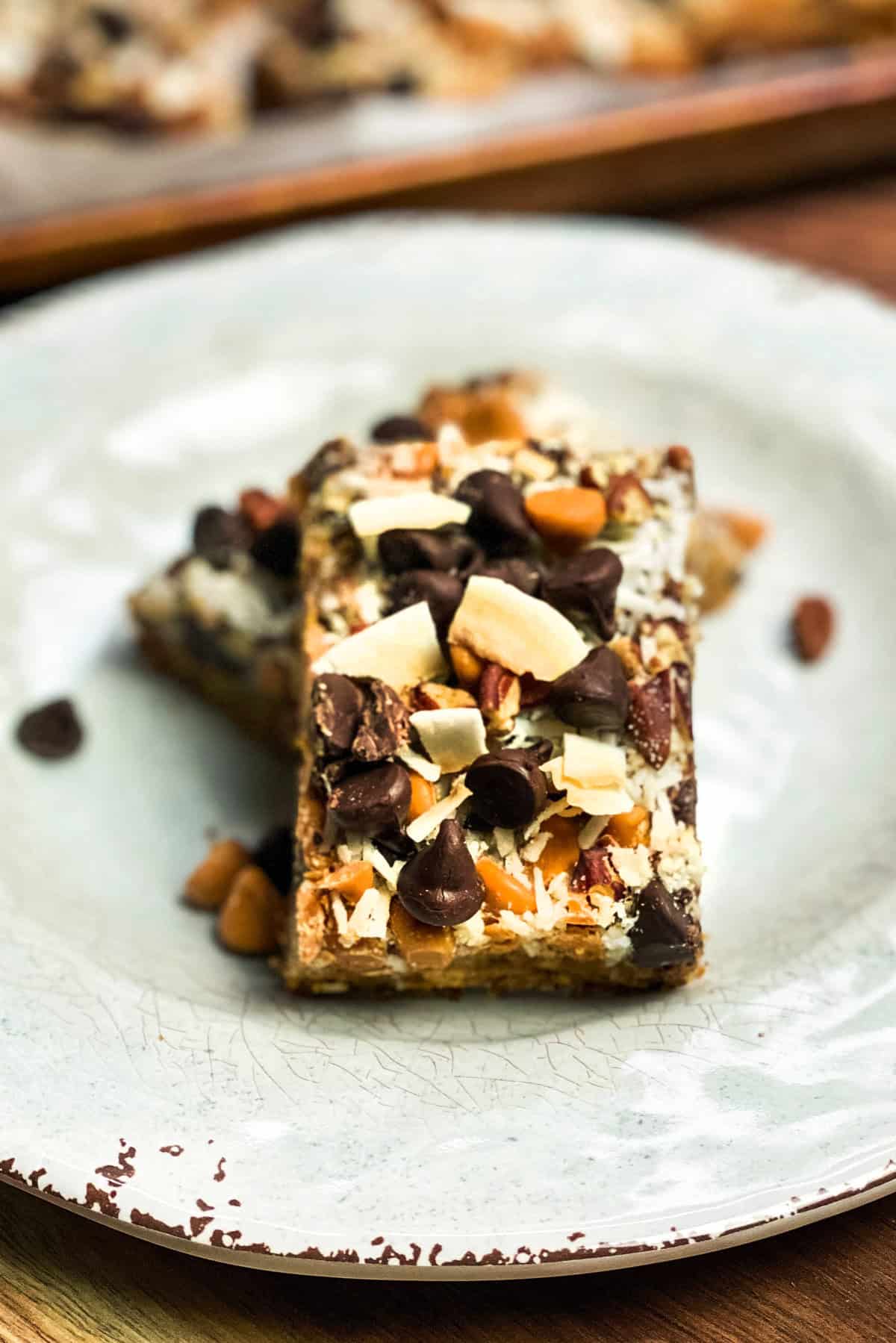 Two Magic Cookie Bars rest enticingly on a white plate, adorned with chocolate chips, nuts, and coconut flakes, all beautifully presented on a rustic wooden table.