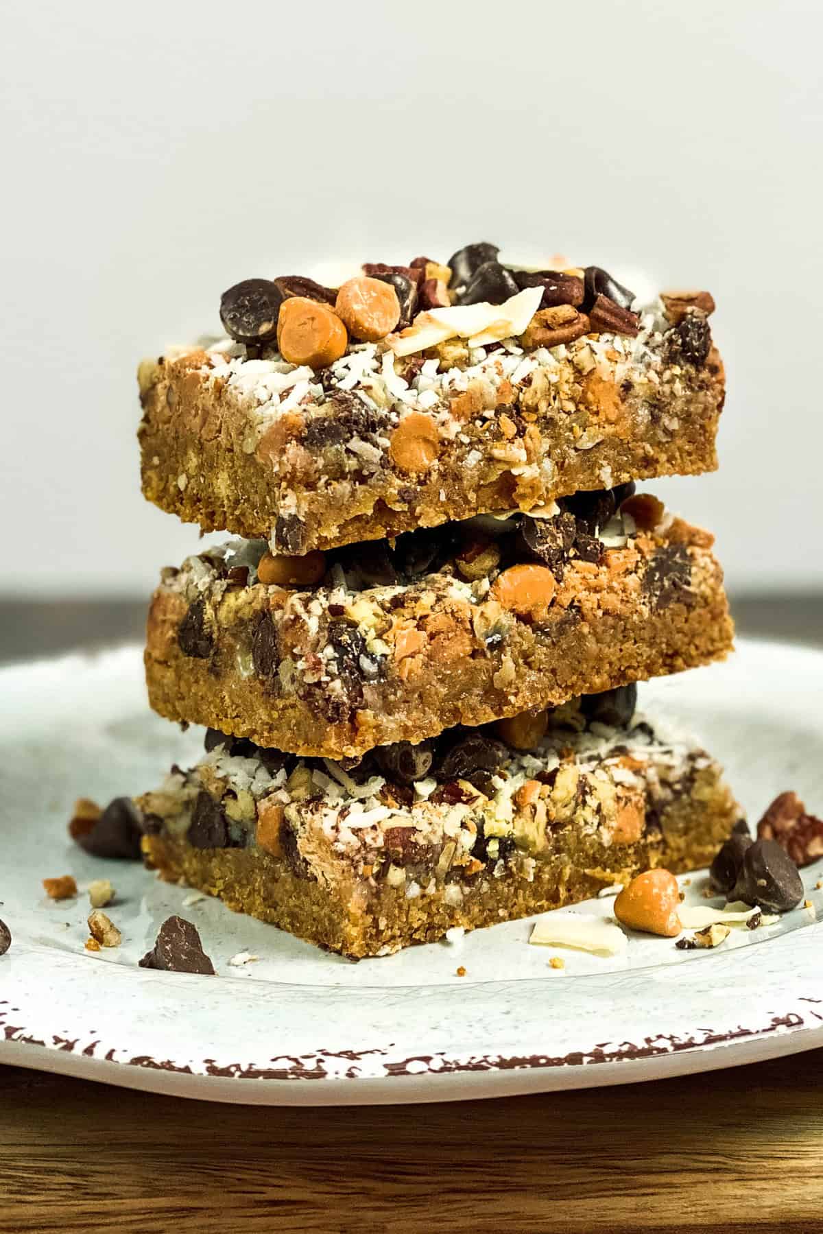 A stack of three Magic Cookie Bars sits on a parchment-lined plate. The bars are topped with chocolate chips, butterscotch chips, and shredded coconut, with visible layers of nuts and a crumbly base. The background is a neutral color.