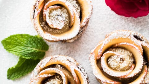 Three apple rose pastries dusted with powdered sugar on a white plate, garnished with mint leaves and two pink roses—perfect for a romantic Valentine's Day breakfast.