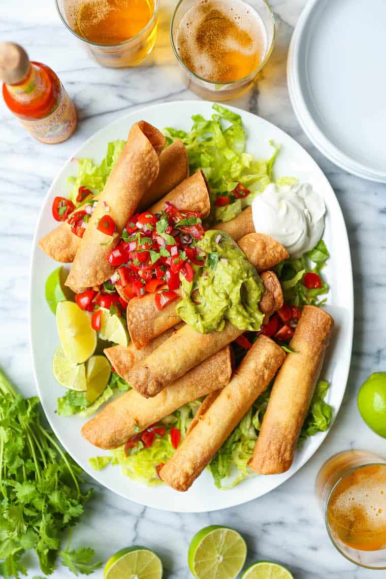 A plate of crispy taquitos topped with guacamole and salsa, served on lettuce with lime wedges, sour cream, and beverages on the side-perfect Super Bowl Party Snacks for your next big game gathering.