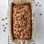 Freshly baked chocolate chip banana bread sits in a parchment-lined loaf pan on a wire rack, with extra chocolate chips scattered around for an irresistible touch.