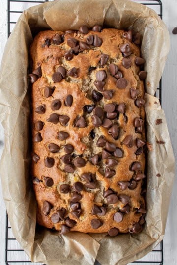 Freshly baked chocolate chip banana bread sits in a parchment-lined loaf pan on a wire rack, with extra chocolate chips scattered around for an irresistible touch.