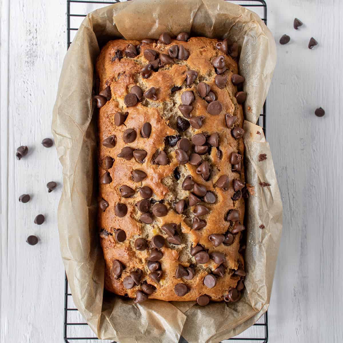 Freshly baked chocolate chip banana bread sits in a parchment-lined loaf pan on a wire rack, with extra chocolate chips scattered around for an irresistible touch.