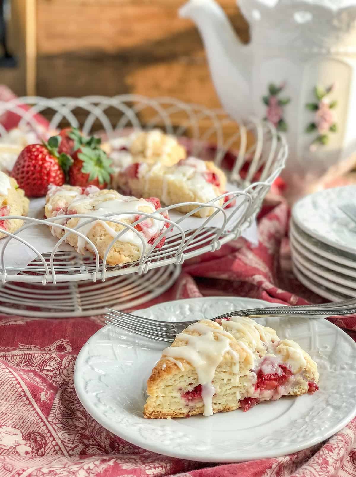 A glazed strawberry scone on a white plate with a fork, with more strawberry scones and fresh strawberries in a basket behind, next to a teapot and stacked plates on a red patterned cloth.