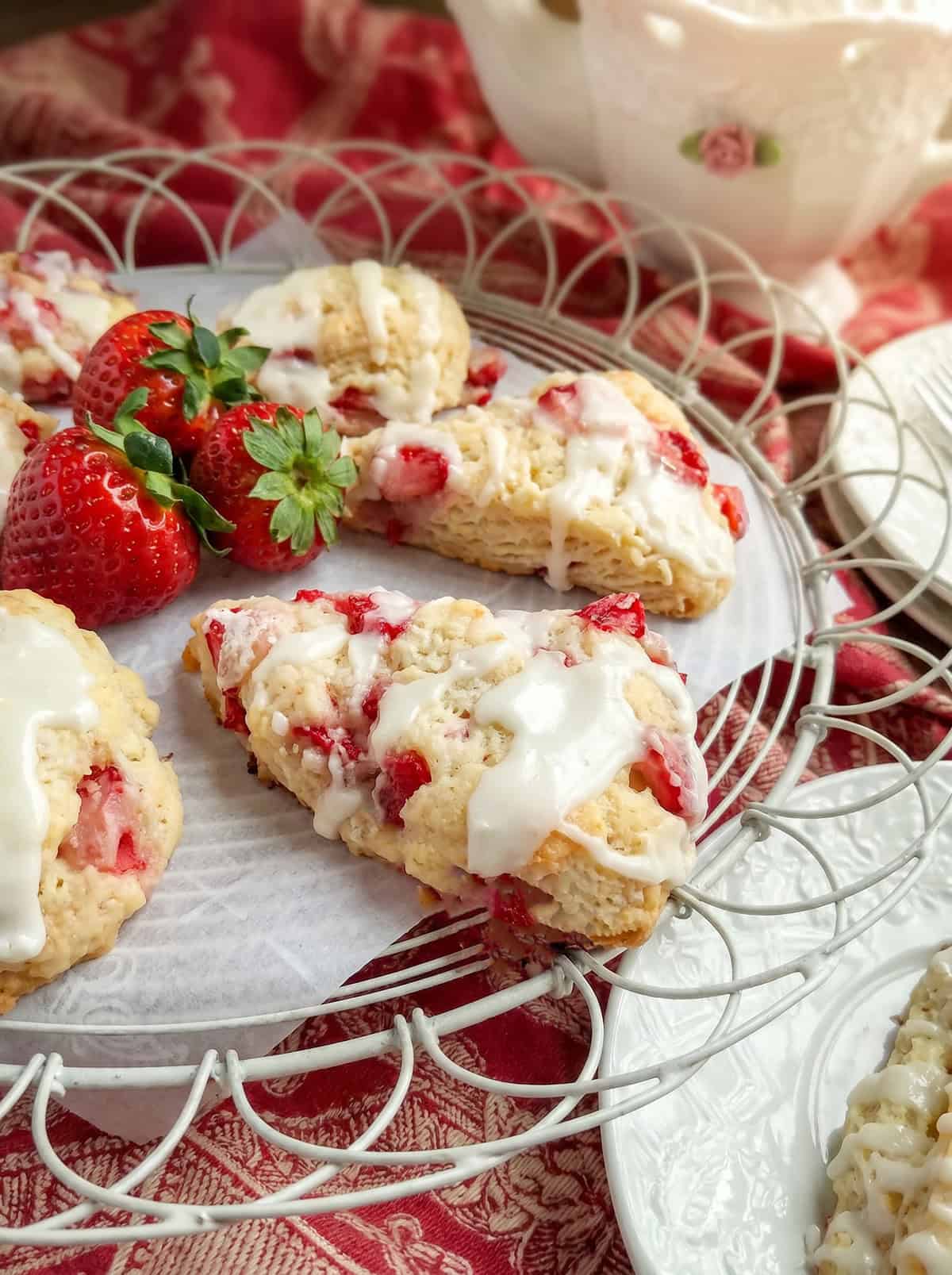 Fresh strawberry scones with white icing are artfully arranged on a wire tray with ripe strawberries, set on a red patterned cloth beside a teacup.