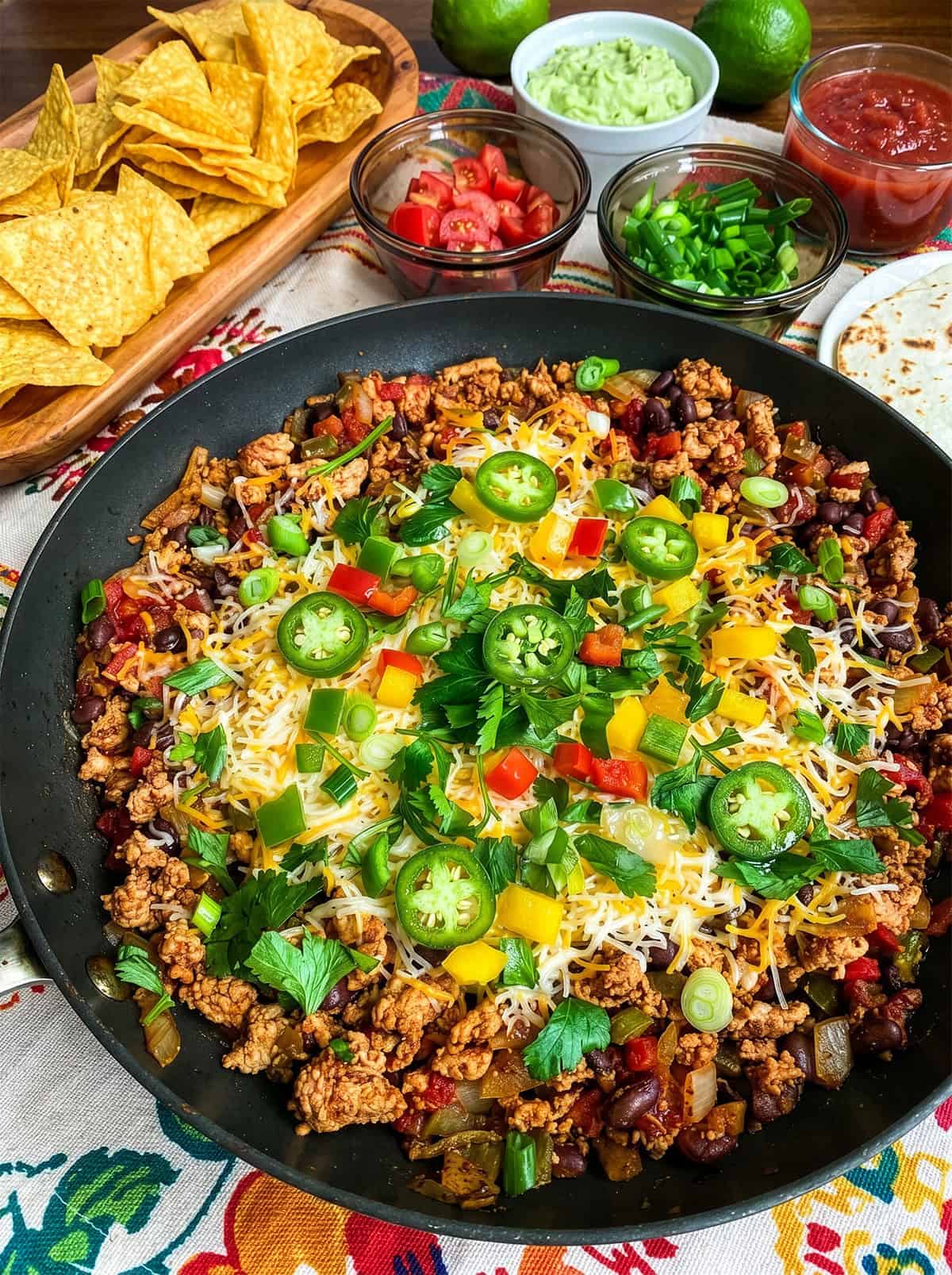 A Skillet Taco dish of seasoned ground meat, beans, and vegetables topped with cheese and sliced jalapeños, surrounded by bowls of tortilla chips, chopped tomatoes, green onions, salsa, and guacamole.