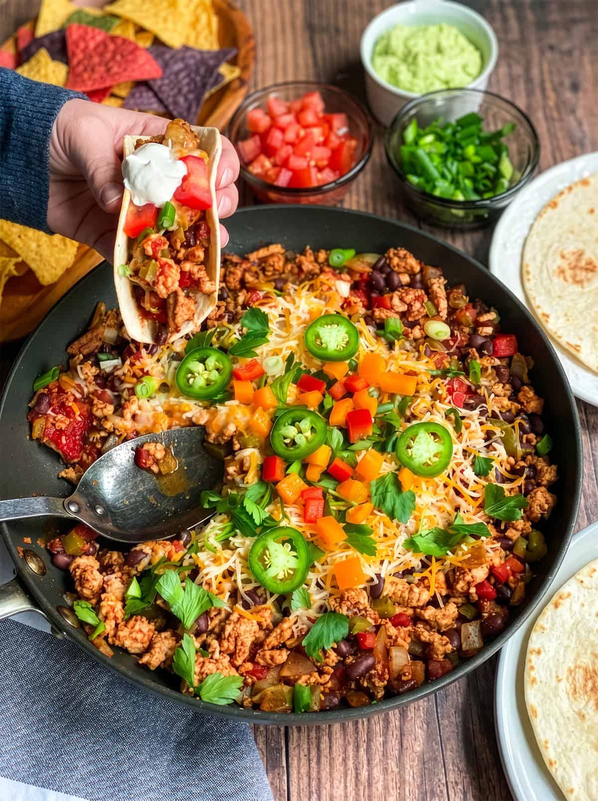 A hand holds a Skillet Taco with sour cream over a skillet filled with seasoned ground meat, beans, cheese, and vegetables, surrounded by taco toppings and tortillas.