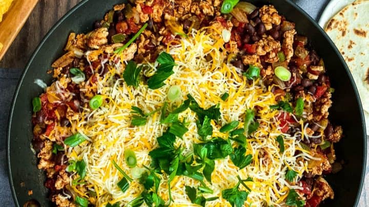 A skillet filled with seasoned ground meat, tomatoes, onions, and melted shredded cheese, topped with green onions and cilantro—an ideal January One Pot Dinner—served with tortilla chips, chopped tomatoes, guacamole, salsa, and tortillas.