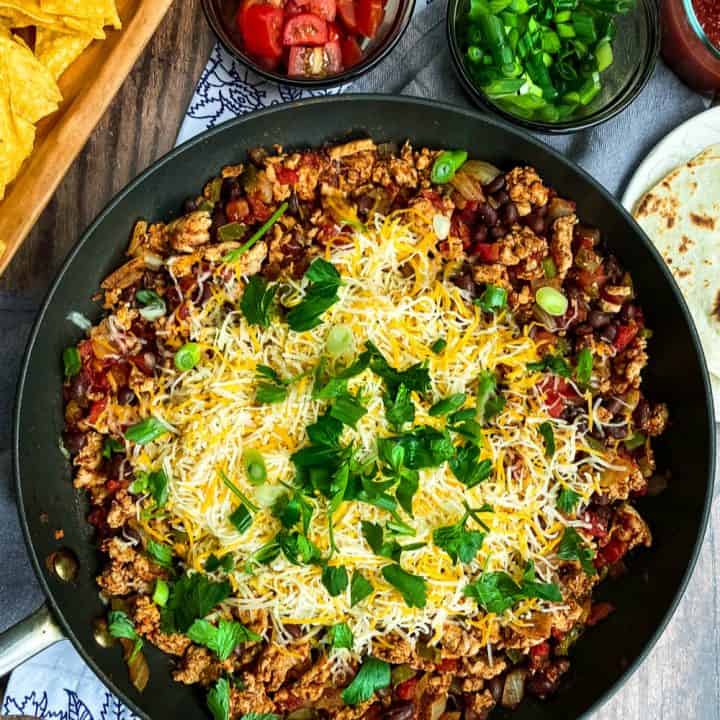 A skillet filled with seasoned ground meat, tomatoes, onions, and melted shredded cheese, topped with green onions and cilantro—an ideal January One Pot Dinner—served with tortilla chips, chopped tomatoes, guacamole, salsa, and tortillas.