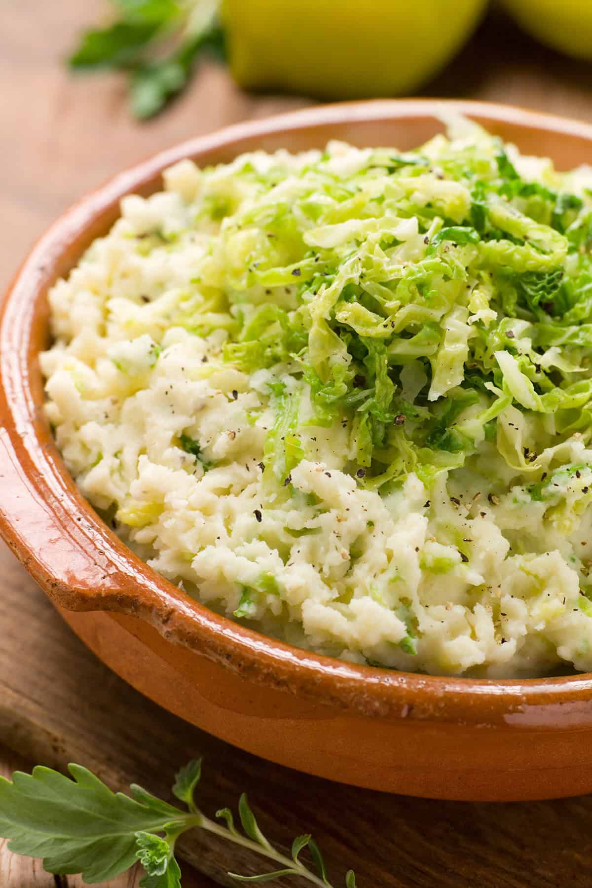 A bowl of Colcannon—mashed potatoes topped with shredded green cabbage—served in a brown ceramic dish on a wooden surface.