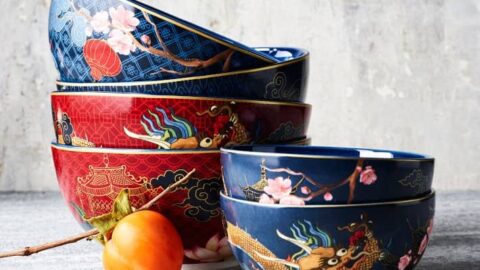 A stack of four decorative bowls with dragon and cherry blossom designs, perfect for celebrating Lunar Chinese New Year, sits next to an orange persimmon fruit on a gray surface.