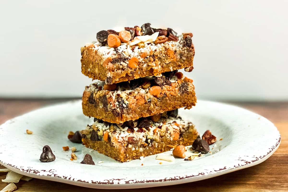 A stack of three Magic Cookie Bars sits on a white, textured plate. These delectable bars are topped with chocolate chips, butterscotch chips, and nuts. The backdrop features a simple wooden surface and a plain white wall, enhancing their delicious appeal.