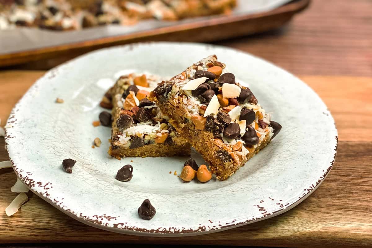 Two Magic Cookie Bars with chocolate chips, nuts, and butterscotch pieces sit stacked on a rustic white plate. A baking tray with more bars is in the background, set against a wooden surface.