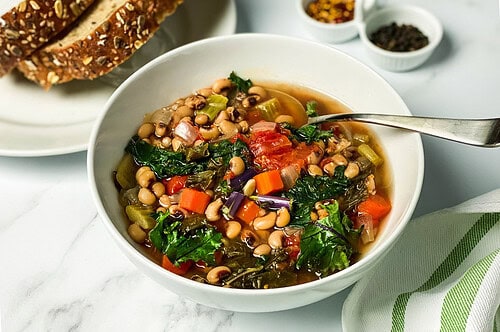 A bowl of hearty vegetable and bean soup, reminiscent of an Ikarian stew, brims with kale, carrots, celery, tomatoes, and black-eyed peas. A spoon rests in the bowl while a plate beside it holds slices of multigrain bread. Small bowls with spices are in the background.