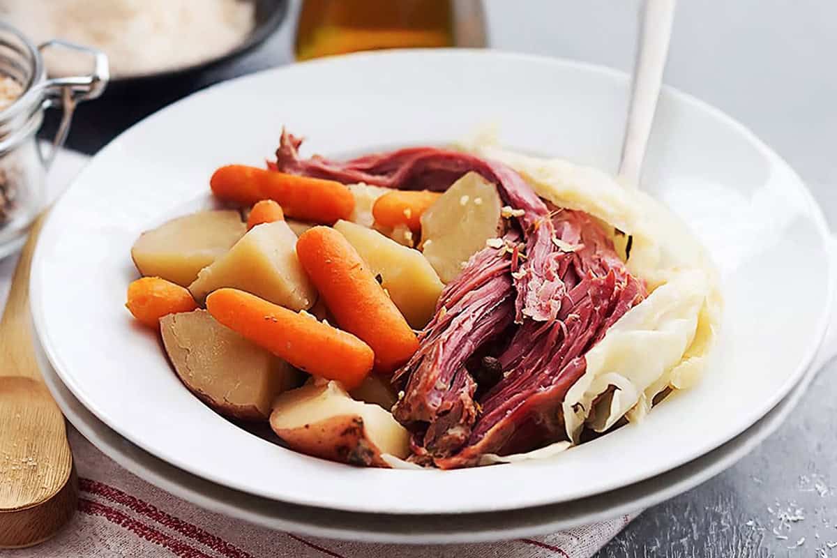 A white plate with Corned Beef and Cabbage, baby carrots, and potatoes, with a fork resting on the side of the plate.