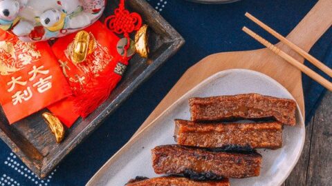 A plate of sliced fried nian gao, a round pan of steamed nian gao with dates, red envelopes, and gold chocolate on a wooden table celebrate Lunar Chinese New Year traditions.