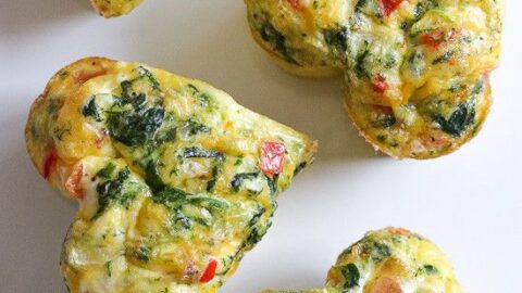 Heart-shaped egg muffins with visible pieces of spinach, red bell pepper, and tomato make a perfect Valentine's Day breakfast on a white surface.