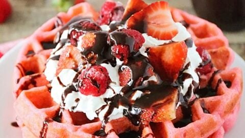 A stack of pink waffles topped with whipped cream, strawberries, raspberries, and chocolate sauce, served on a white plate with a red checked napkin—perfect for a sweet Valentine's Day breakfast.