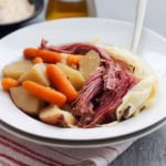 A white bowl filled with classic Corned Beef and Cabbage, potatoes, and baby carrots, served on a red-striped cloth.