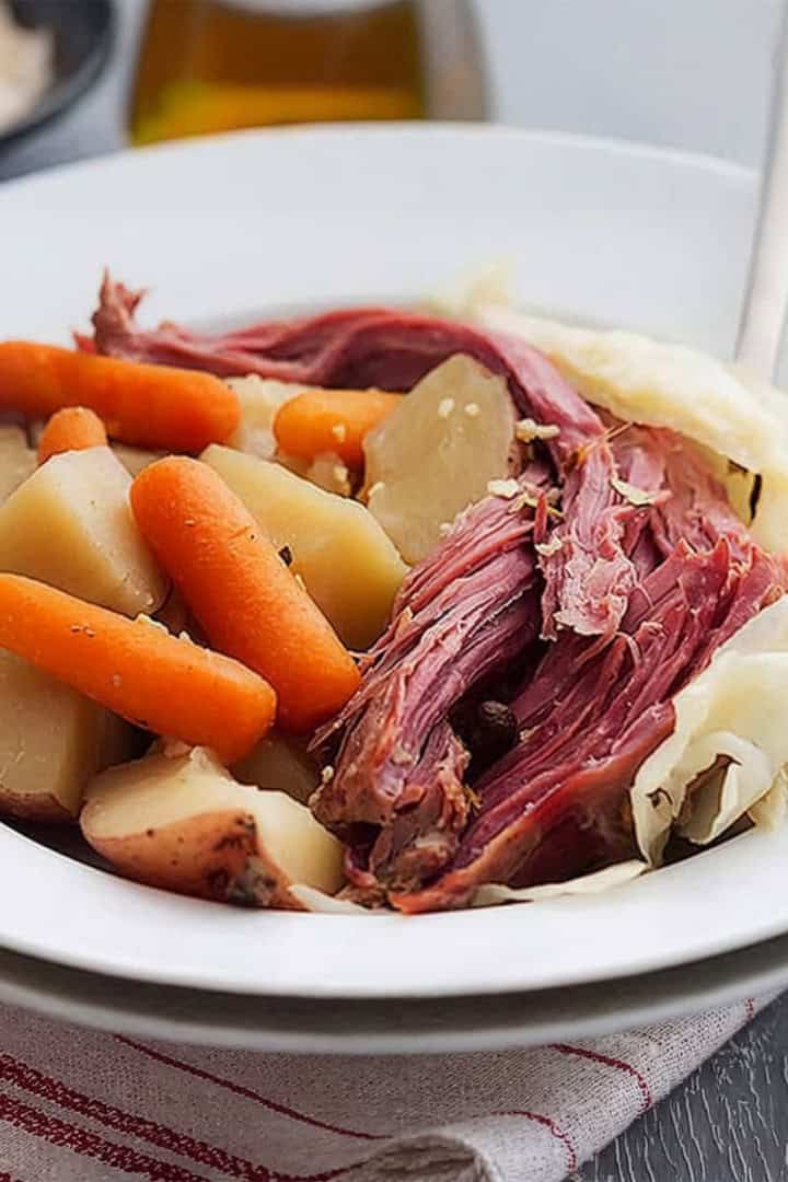 A white bowl filled with classic Corned Beef and Cabbage, potatoes, and baby carrots, served on a red-striped cloth.