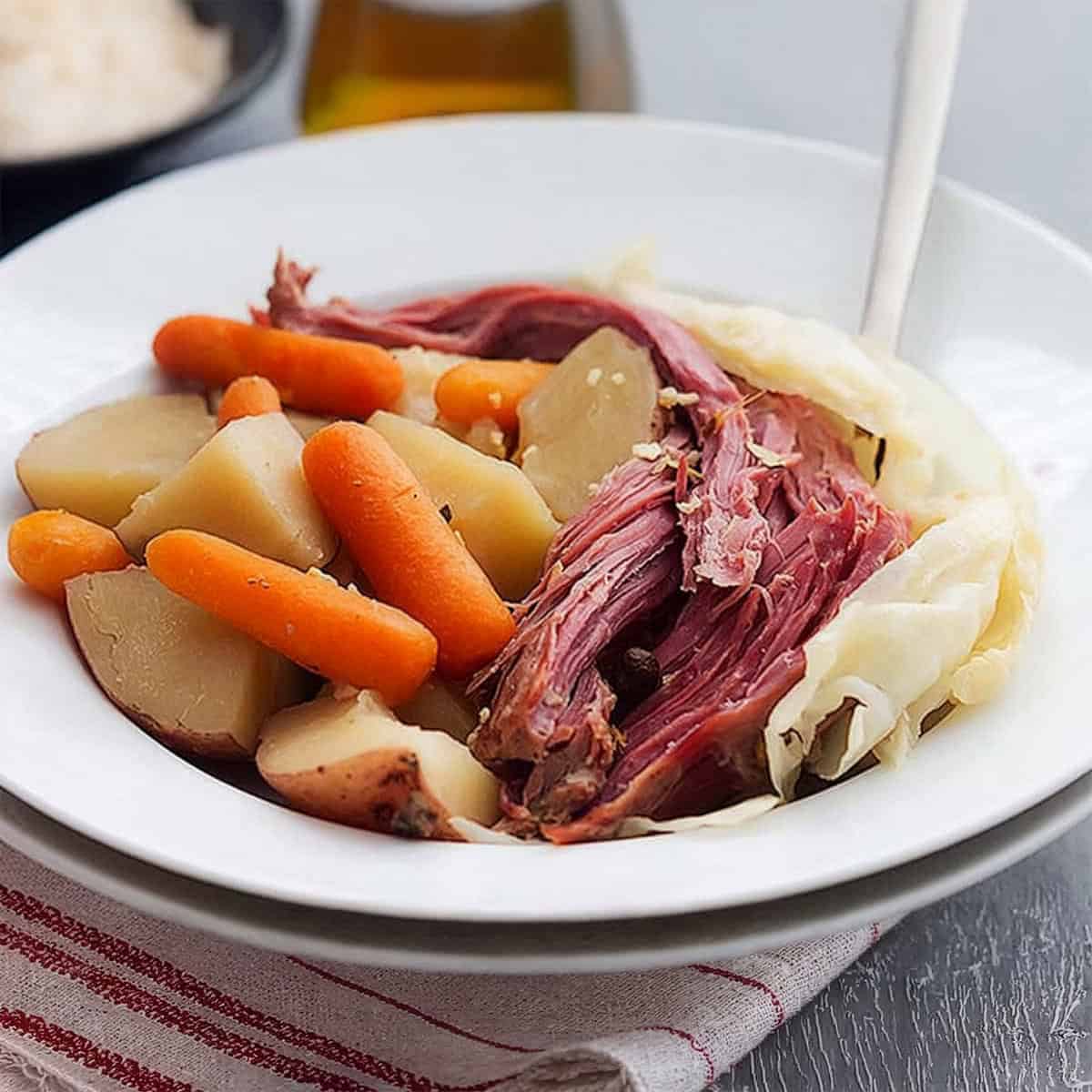 A white bowl filled with classic Corned Beef and Cabbage, potatoes, and baby carrots, served on a red-striped cloth.