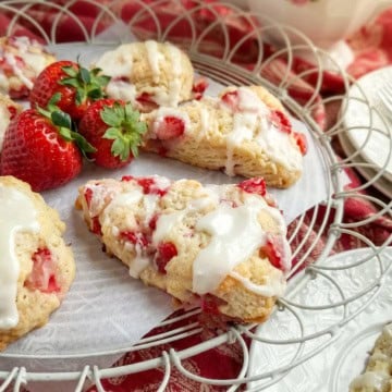 A plate of strawberry scones with white icing, garnished with fresh strawberries, is displayed on a wire tray with a red patterned cloth underneath-perfect for showcasing these delicious strawberry scones.