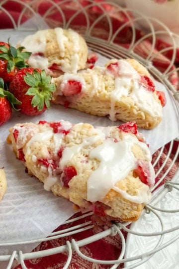 A plate of strawberry scones with white icing, garnished with fresh strawberries, is displayed on a wire tray with a red patterned cloth underneath-perfect for showcasing these delicious strawberry scones.