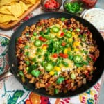 A Skillet Taco with cooked ground meat, beans, melted cheese, diced peppers, green onions, and jalapeño slices in a skillet, surrounded by bowls of chips, tomatoes, salsa, green onions, and tortillas.