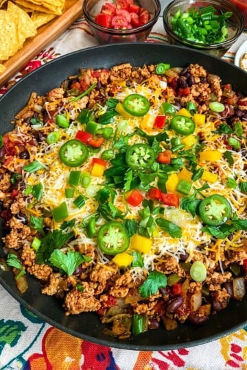 A Skillet Taco with cooked ground meat, beans, melted cheese, diced peppers, green onions, and jalapeño slices in a skillet, surrounded by bowls of chips, tomatoes, salsa, green onions, and tortillas.